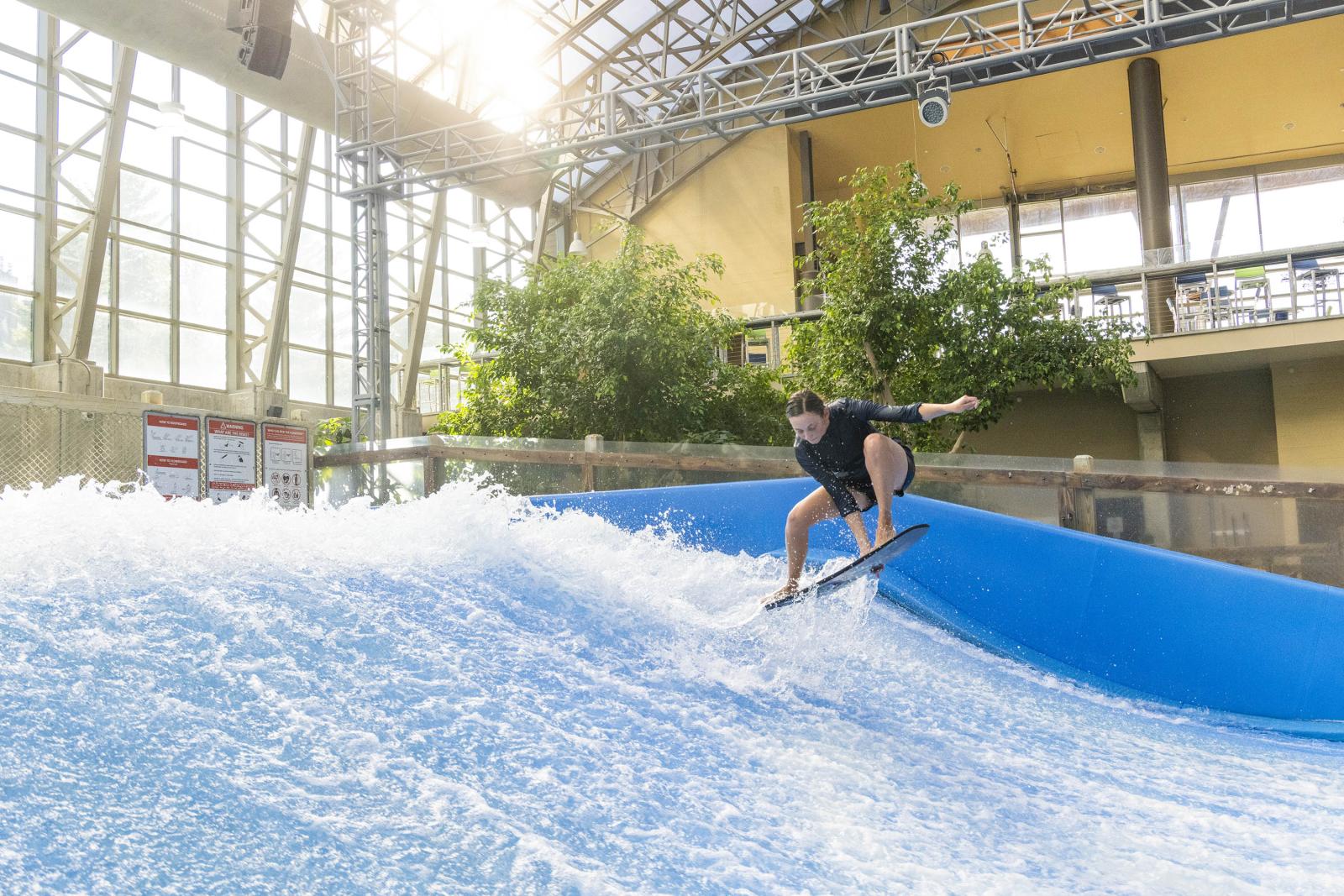 Surfer riding an indoor wave with sunlight streaming through large windows.