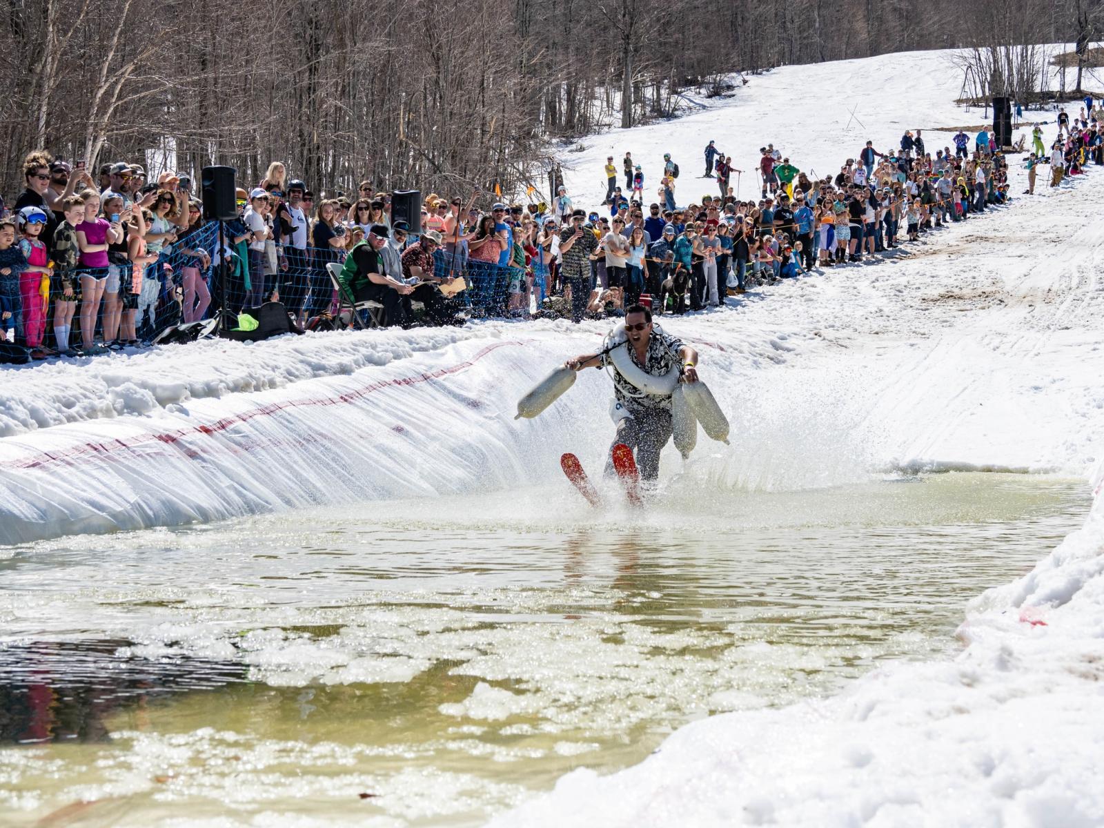Skier skimming a snowy pond, watched by a crowd on a winter day.