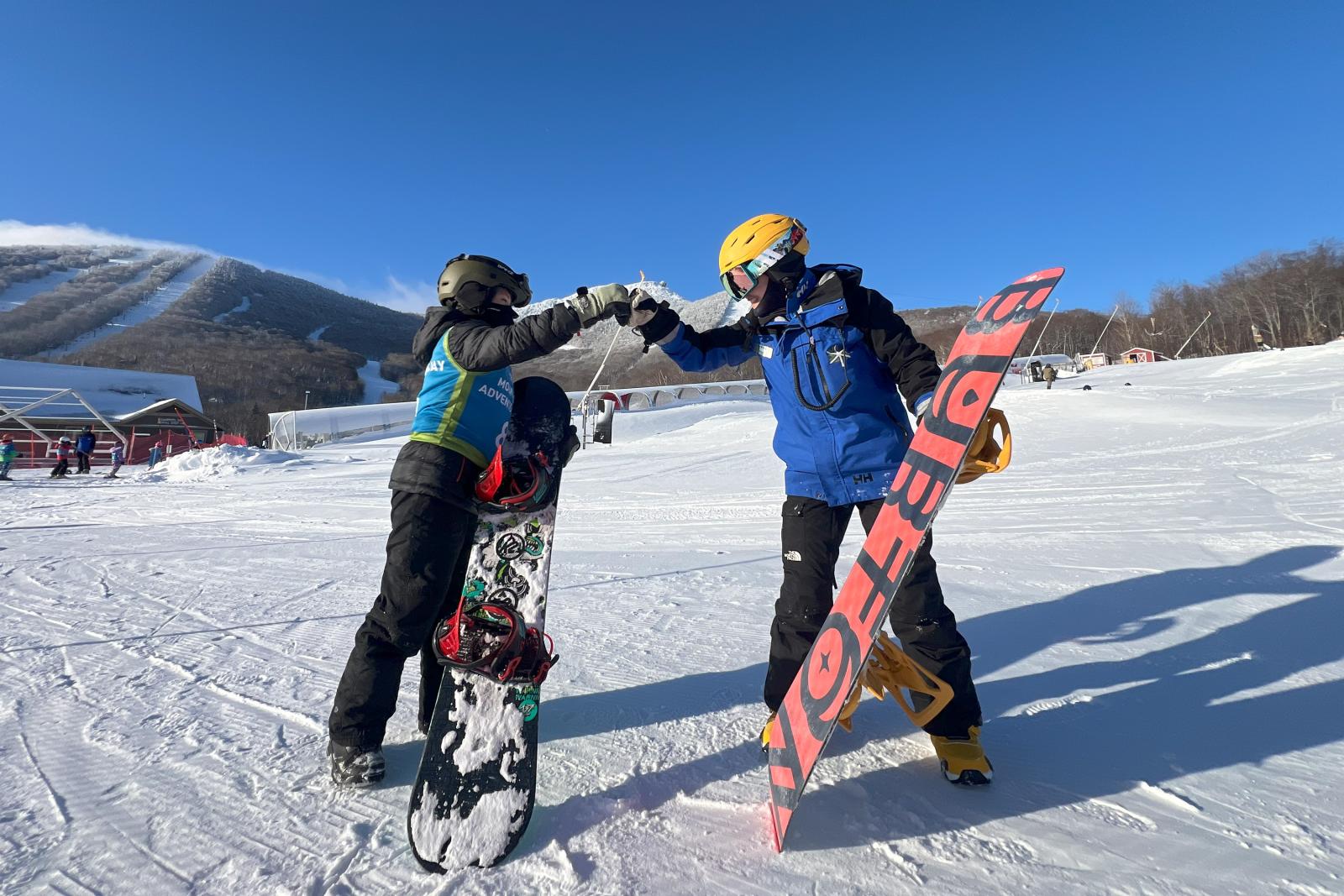 Two snowboarders fist bump on a snowy mountain with clear blue sky.