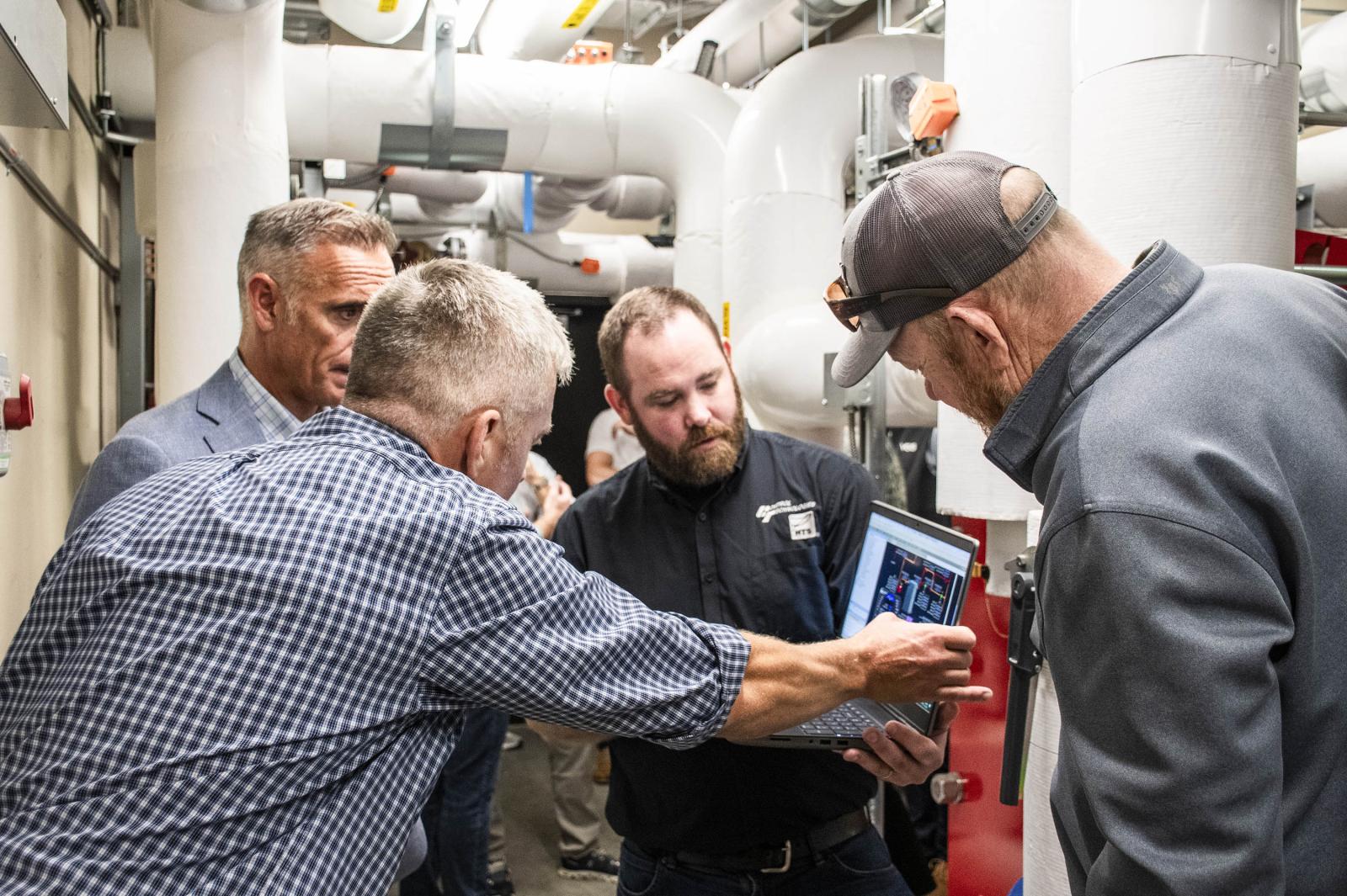 Four men in a mechanical room examining a laptop screen.