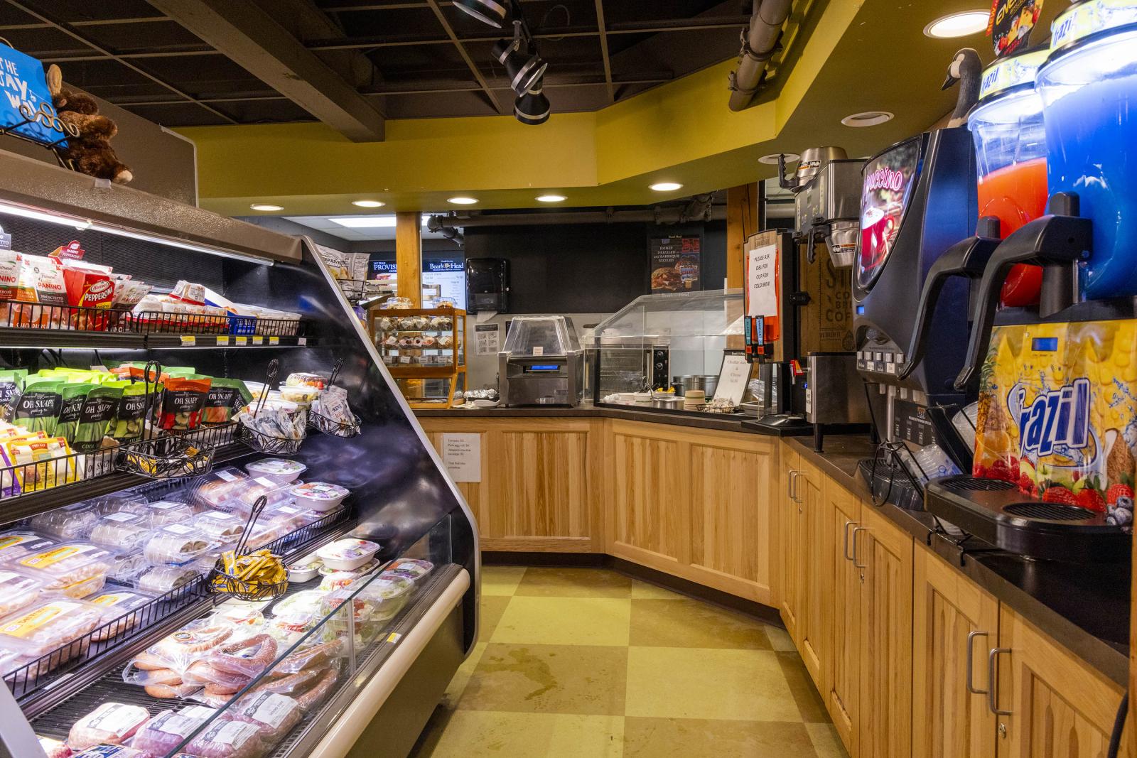 Grocery store deli section, sandwiches and snacks displayed, colorful drinks machine on right.