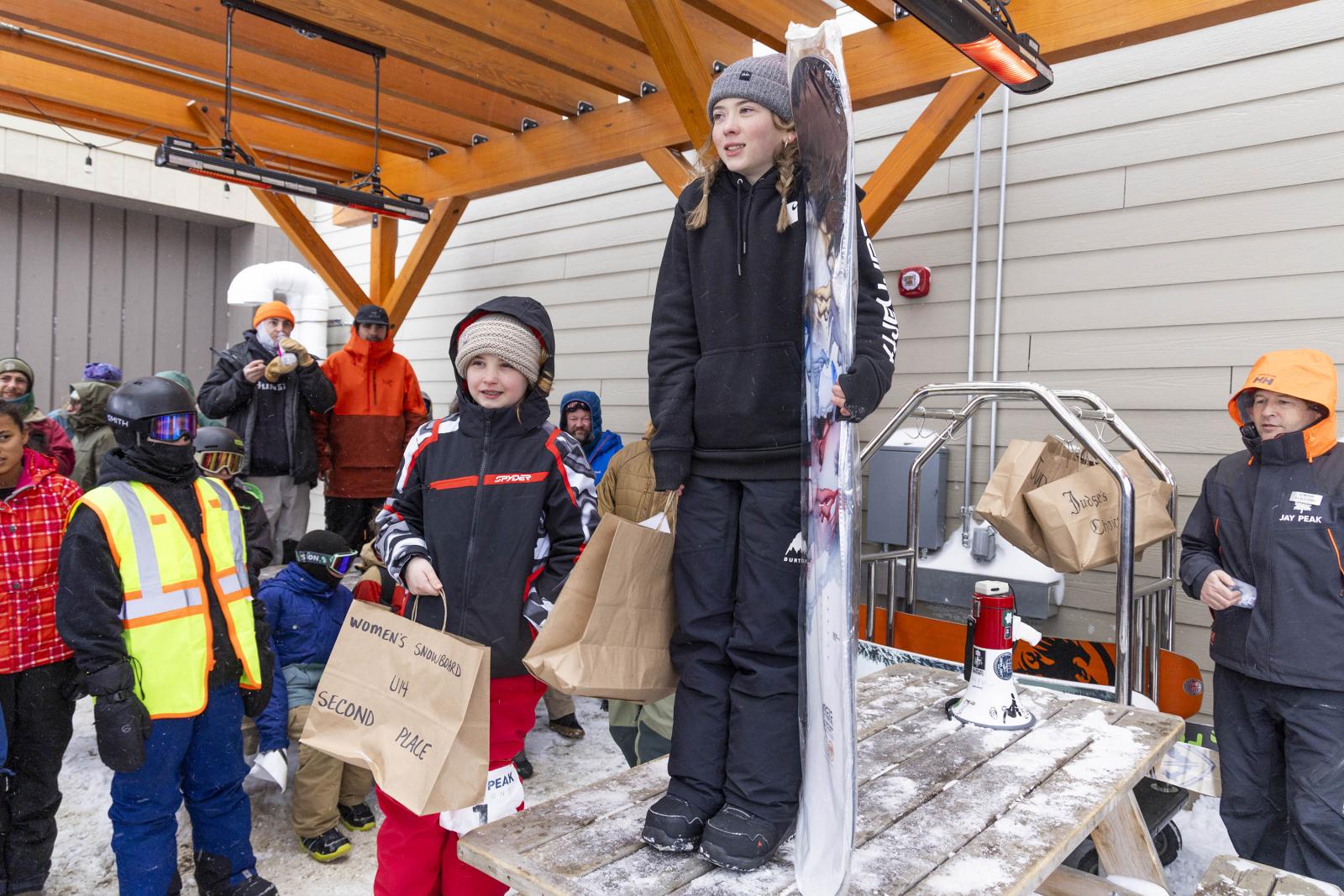 Children holding bags stand on a wooden platform at a winter event.
