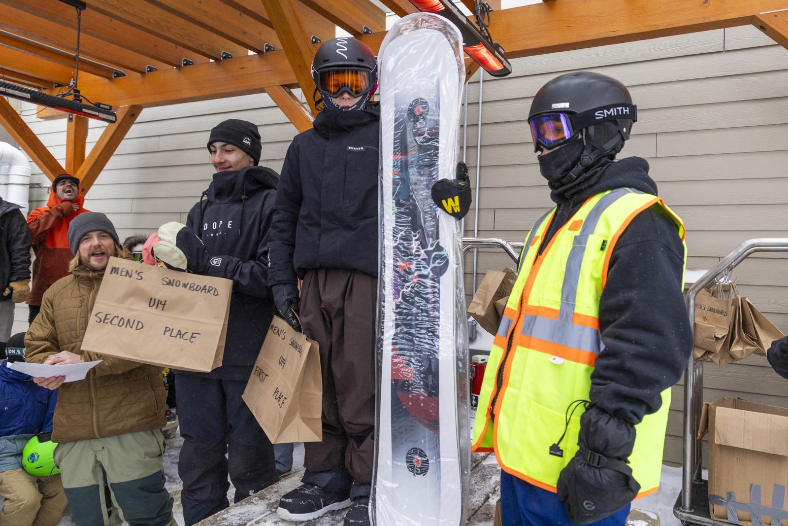 Group of snowboarders in winter gear, one holding a snowboard, outside under wooden shelter.