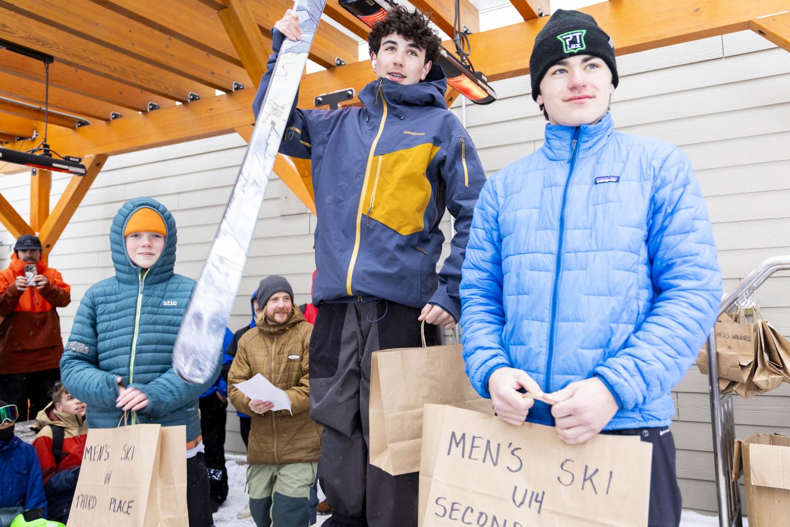 Three people on a podium holding prizes, two in blue jackets, one in a multicolored jacket.