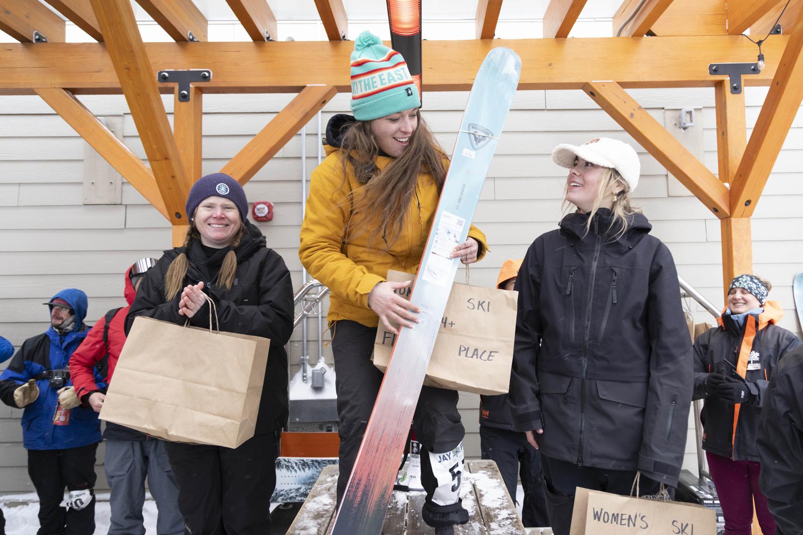 Three women in winter gear holding prizes, one with a snowboard.