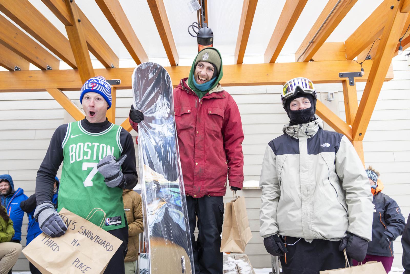 Three snowboarders on a podium, one holding a snowboard, wearing winter gear and smiling.