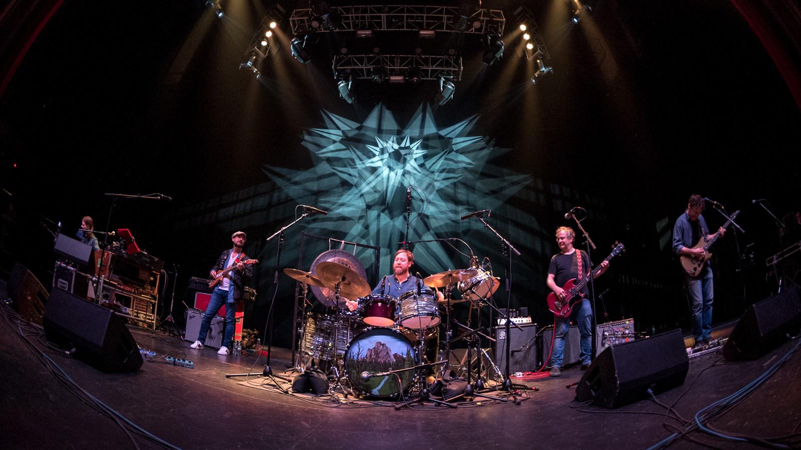 Band performing on stage with a psychedelic background and vibrant lighting.