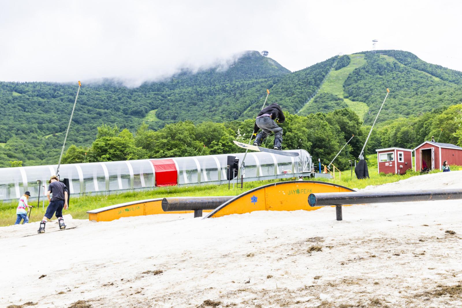 Snowboarder performing a jump on a summer ski slope with green hills in the background.