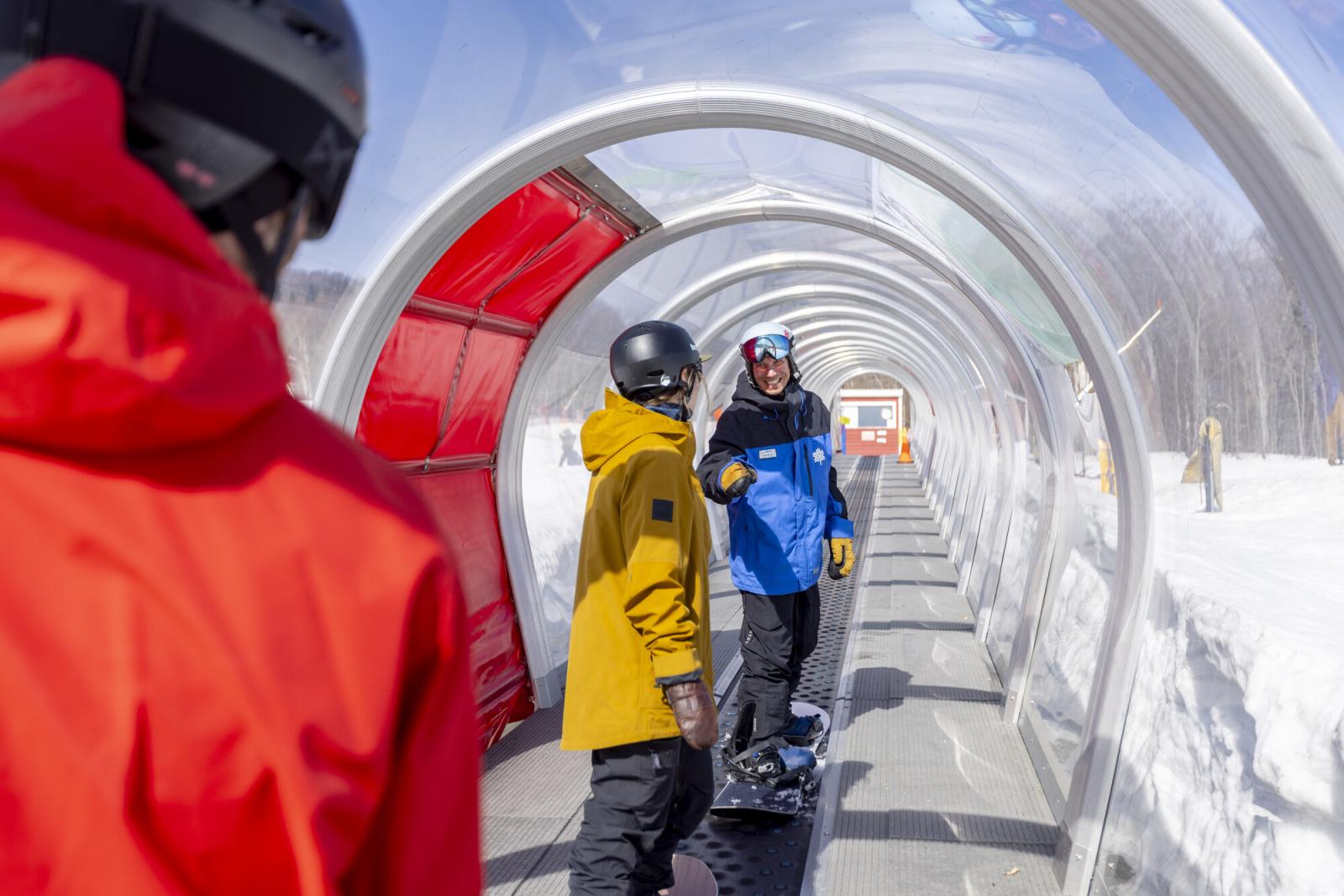 Snowboarders on a covered slope conveyor under clear skies.