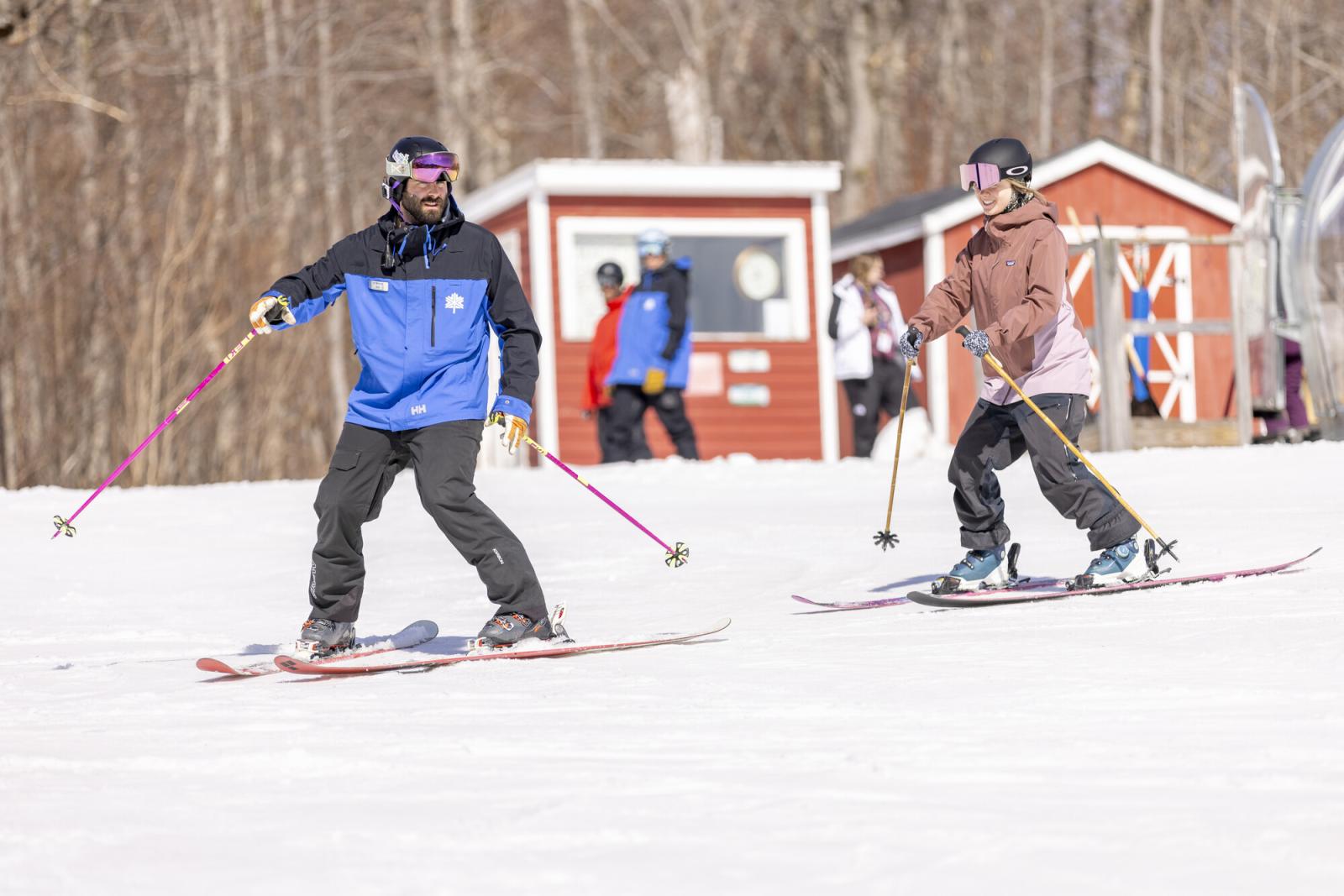 Skiers on a snowy slope with ski poles, trees, and small buildings in the background.