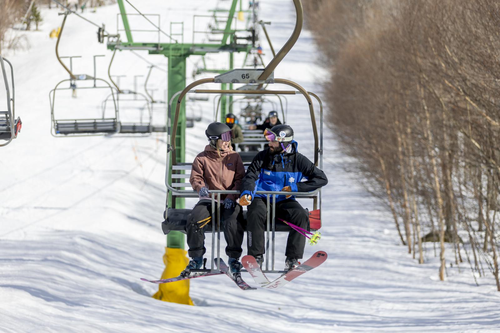 Skiers on a chairlift ascending a snowy slope with bare trees in winter.