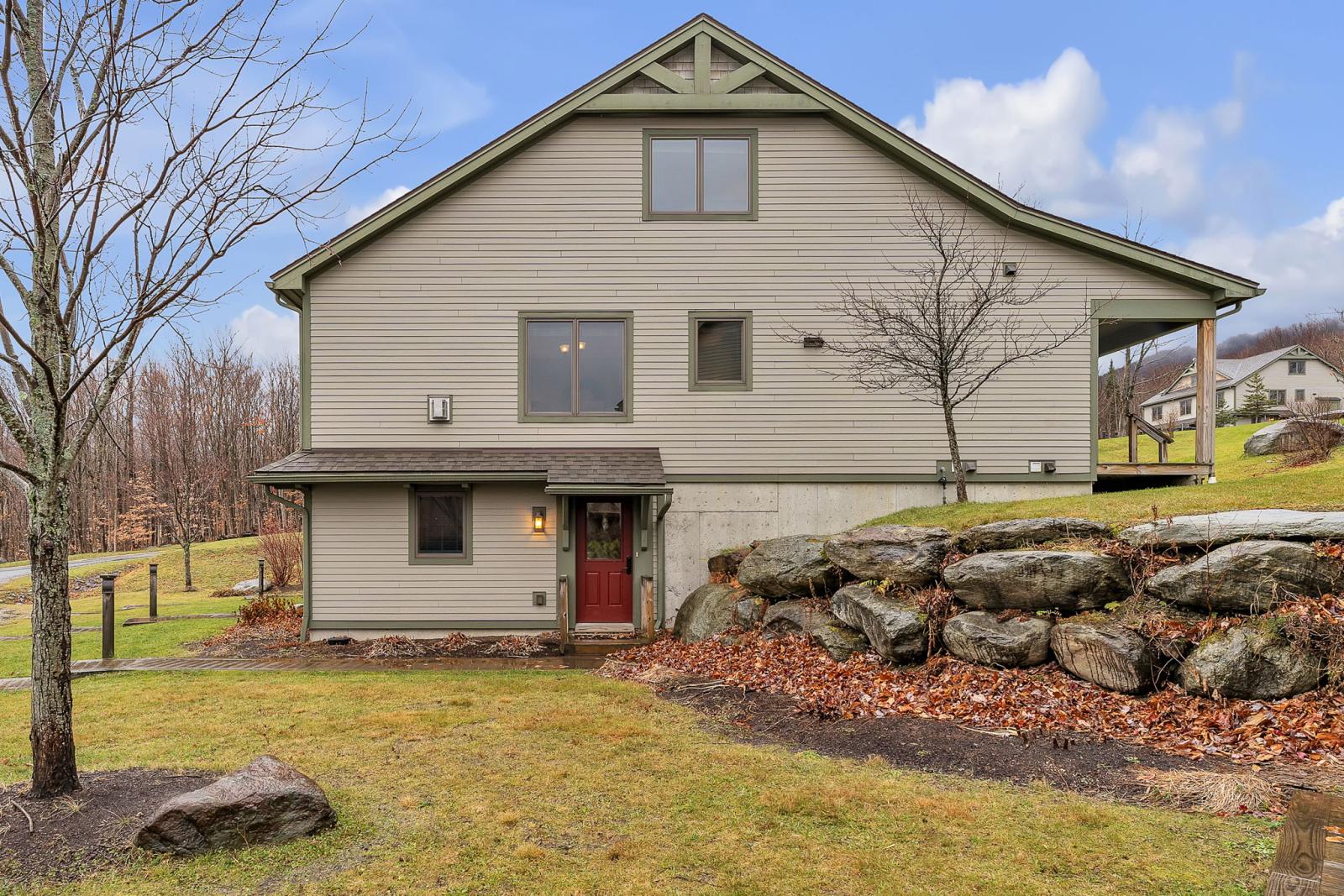 A beige house with red door, surrounded by grass and rocks. Cloudy sky in the background.