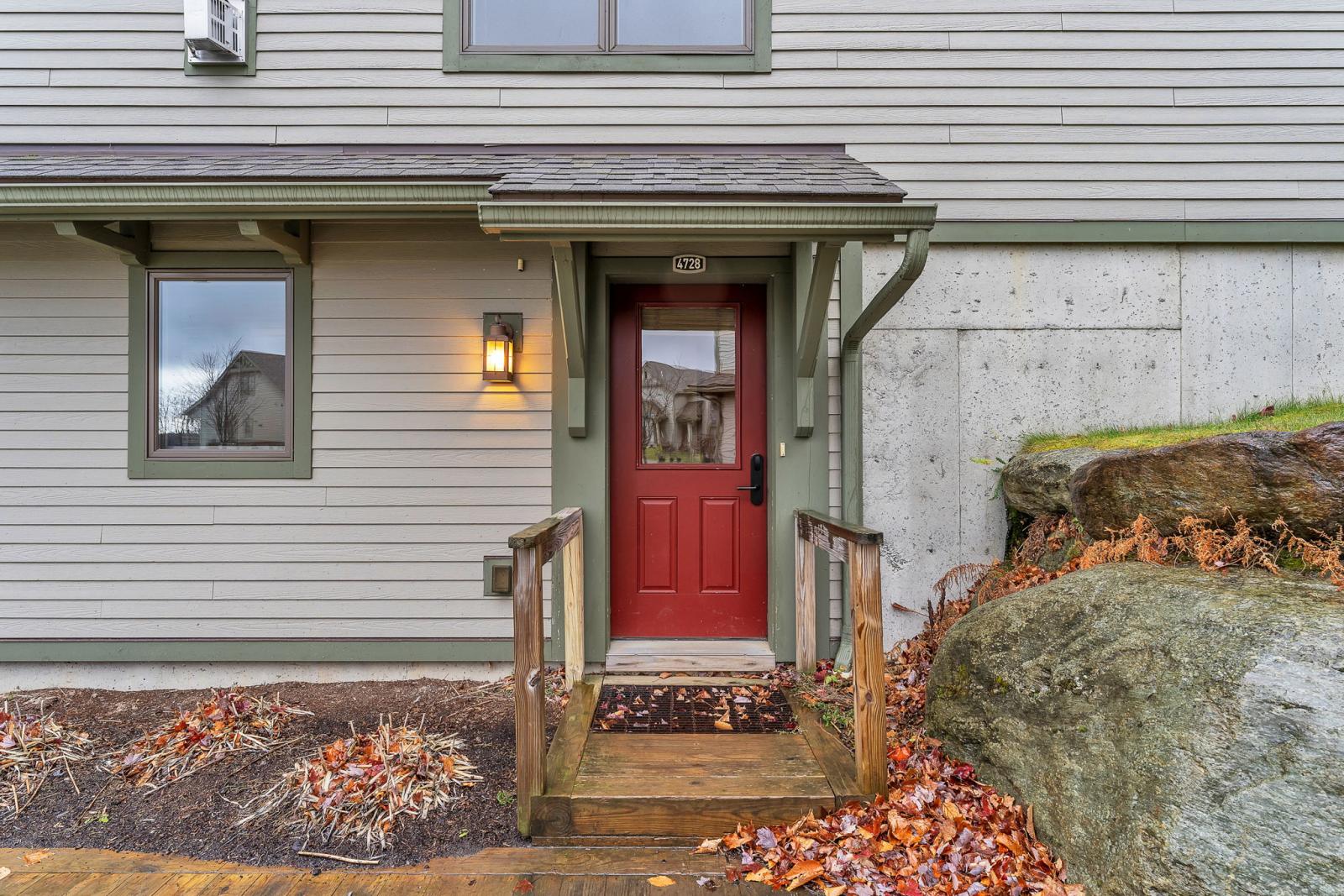 Red door entrance with autumn leaves and a rock beside a wooden ramp.