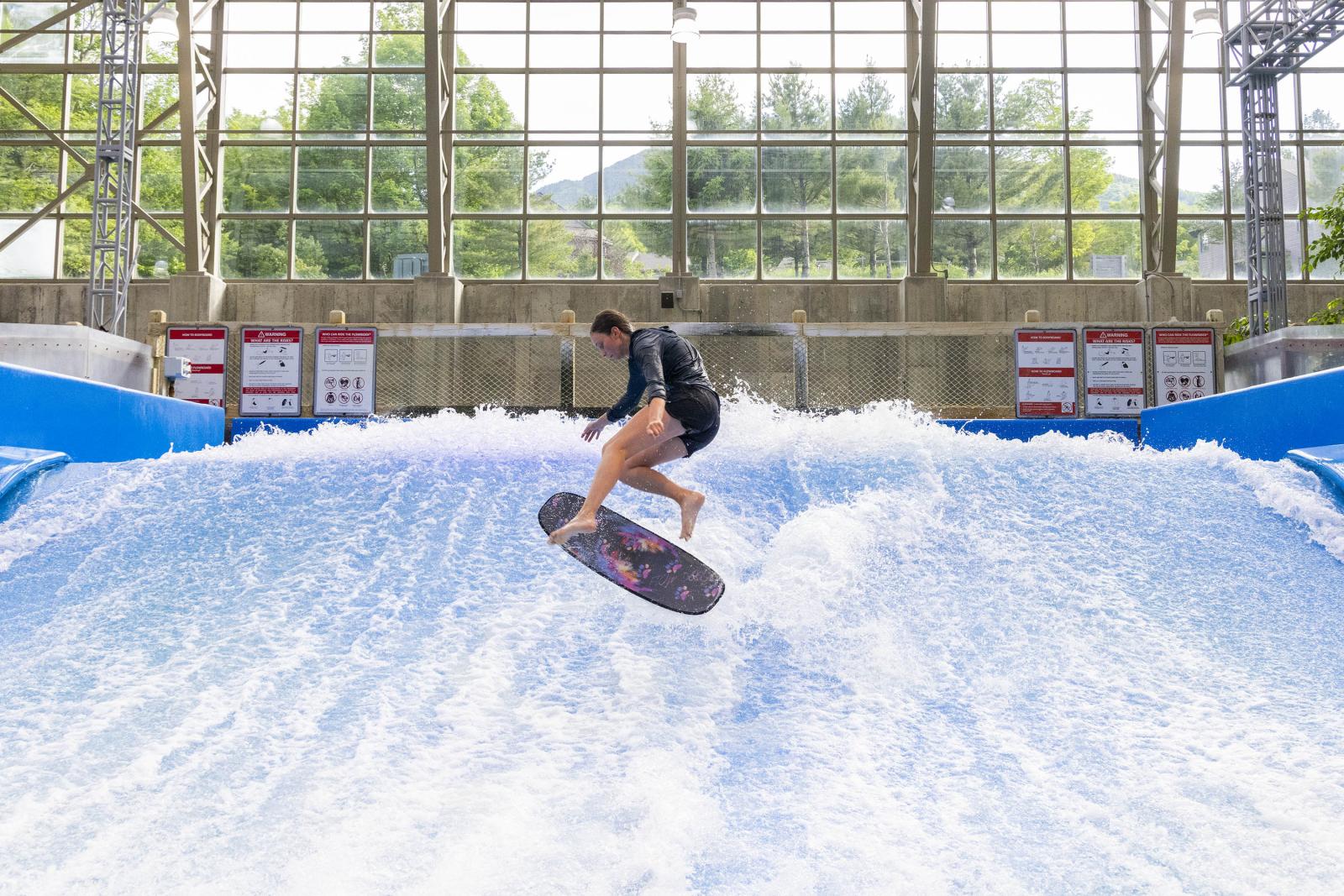 Indoor wave pool with a person surfing, surrounded by windows.