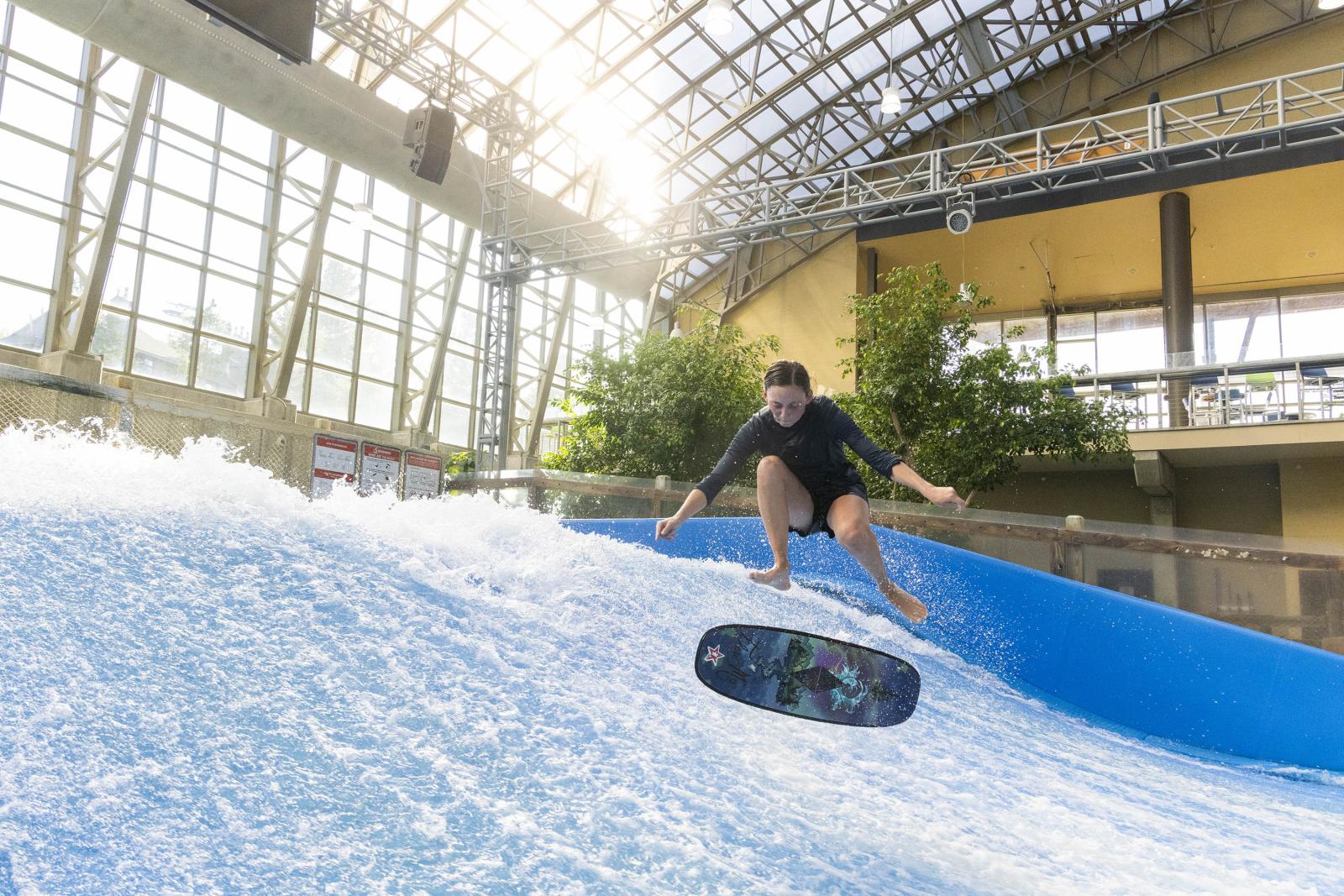 Indoor surfing on artificial wave, person mid-air under glass roof.