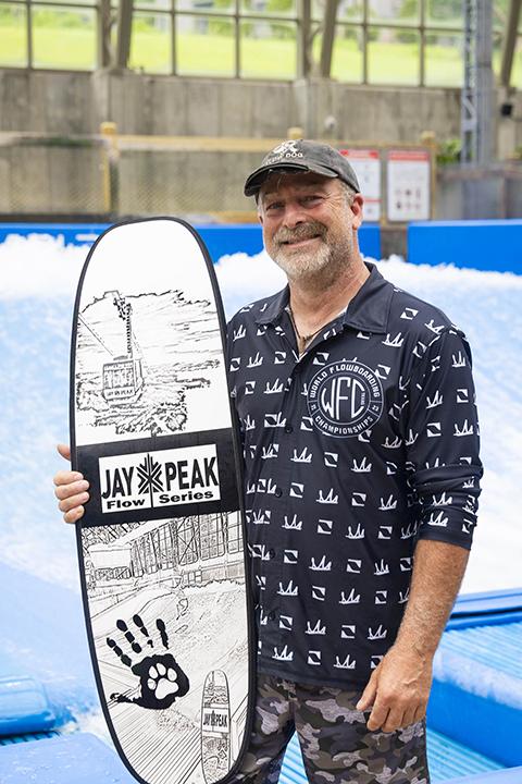 Man holding a skateboard with a smile, wearing a patterned shirt and cap, indoor skate park setting.