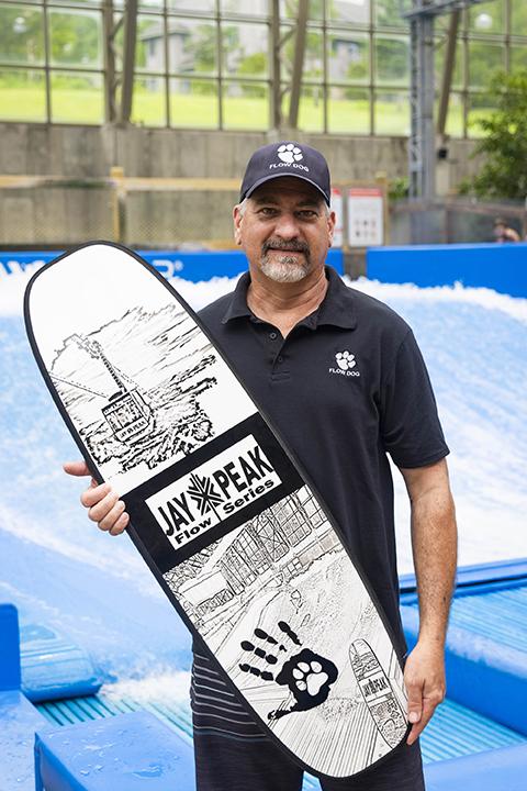 Man in a cap holding a decorated surfboard in a water park setting.