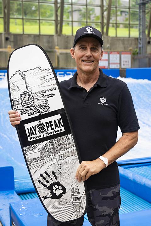 Man in black shirt and cap holding a surfboard indoors.