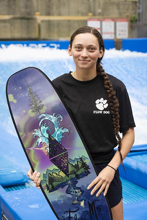 Young woman holding a colorful surfboard, standing by a wave pool.
