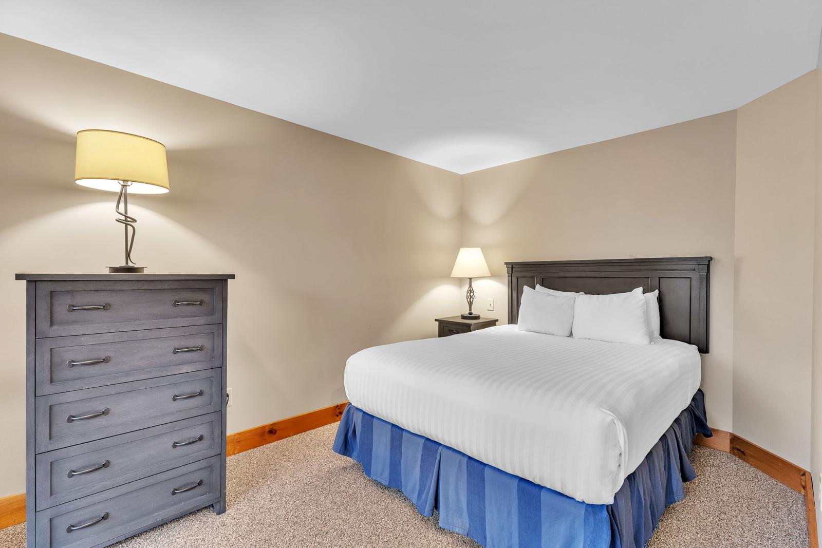 Bedroom with a bed, nightstand, dresser, and two lamps. Neutral tones and blue bed skirt.