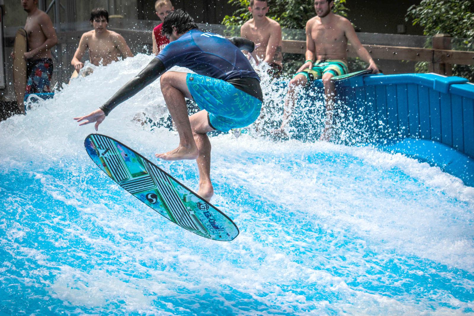 Surfer in blue shorts jumps on artificial wave, spectators watch.