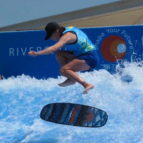 Surfer in mid-air trick on wave pool, wearing a cap and blue vest.