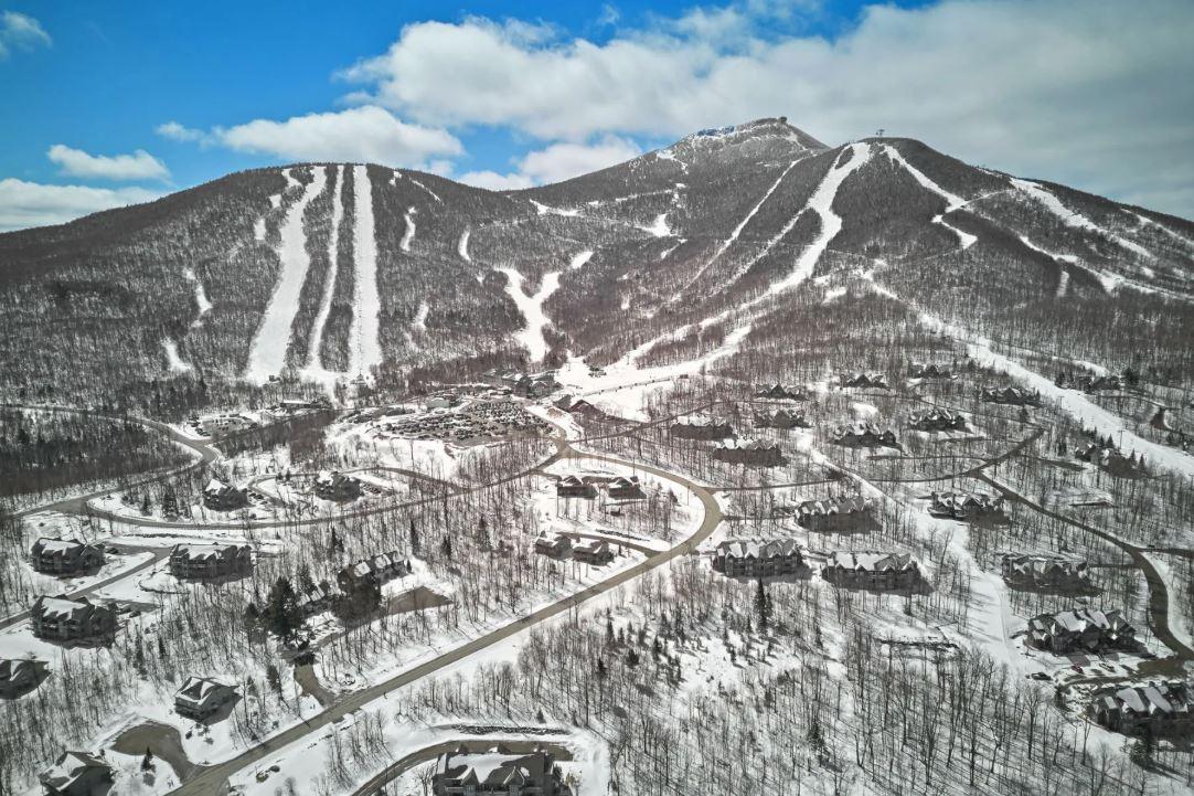 Snow-covered ski resort with slopes, mountains, and scattered houses below a blue sky.