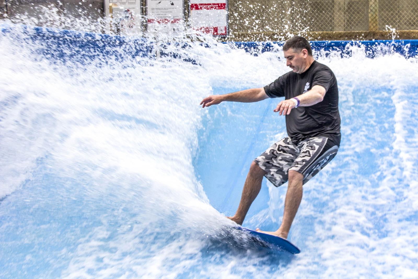 Surfer balancing on an indoor wave, wearing shorts and a black t-shirt.