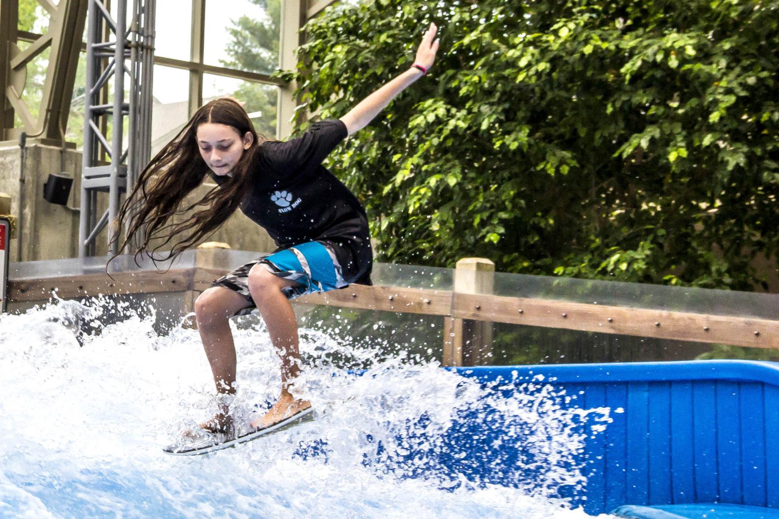 Teen surfing on indoor wave, wearing black shirt and blue shorts, with greenery in background.
