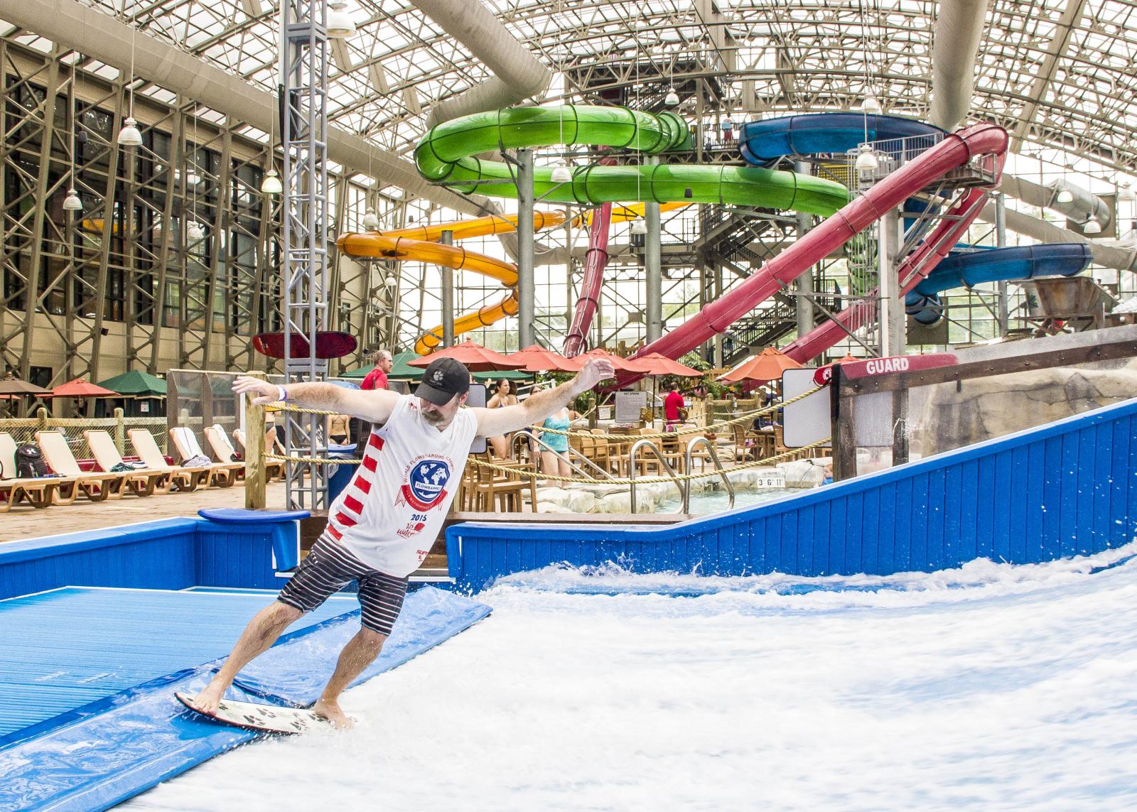 Indoor surfer on artificial wave with colorful water slides in background.