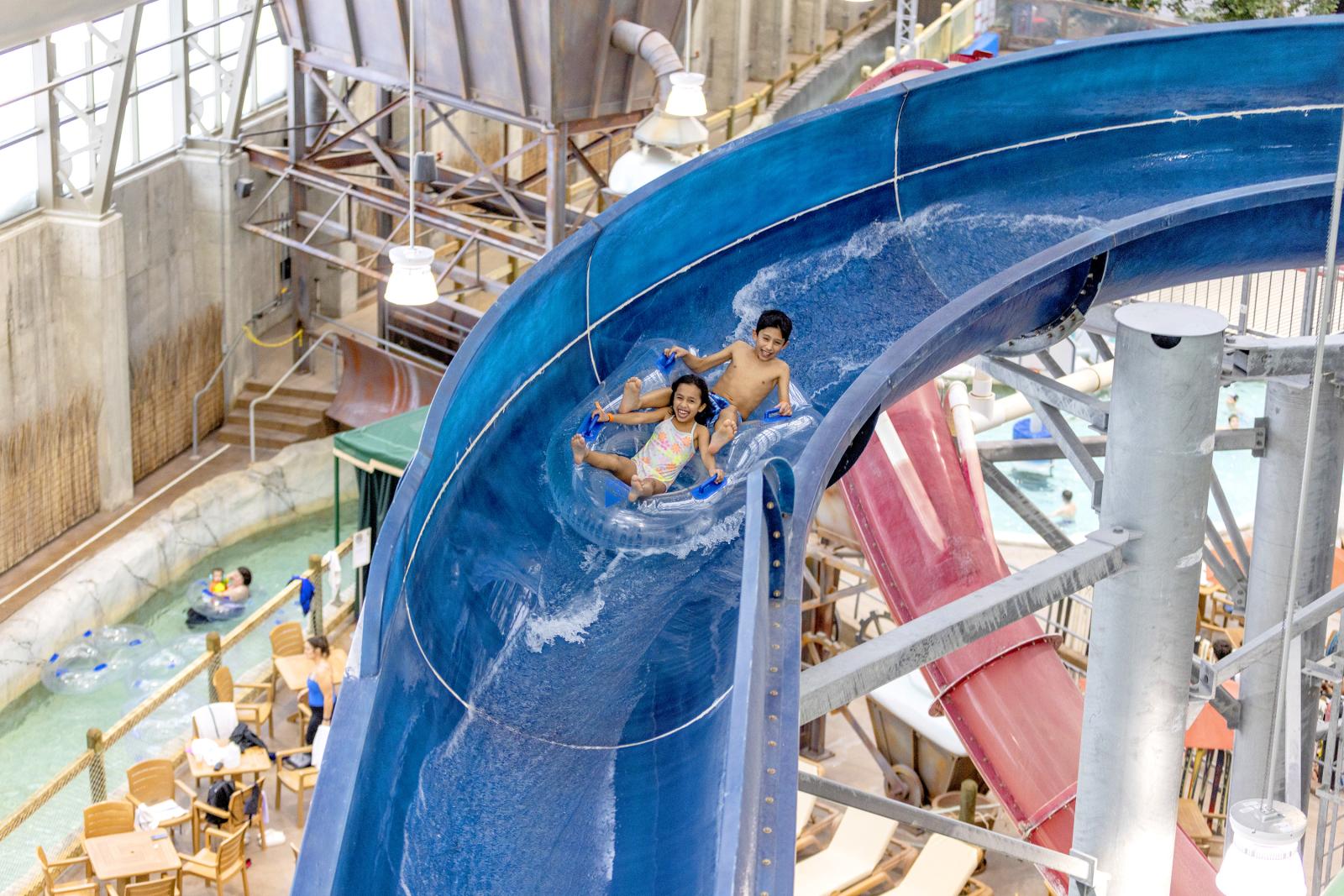 Three people enjoy a ride on a blue water slide indoors.