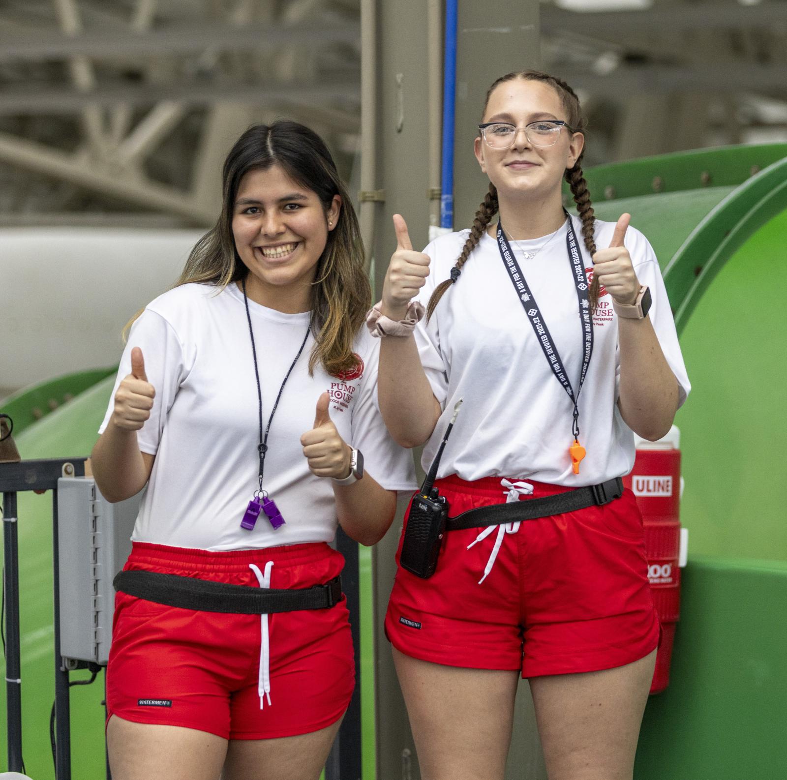 Two lifeguards in red shorts give thumbs up, smiling.