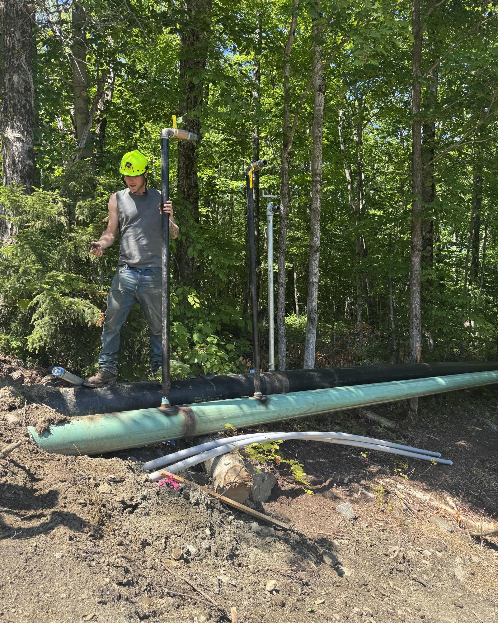 Construction worker with green helmet standing near large pipes in a forested area.