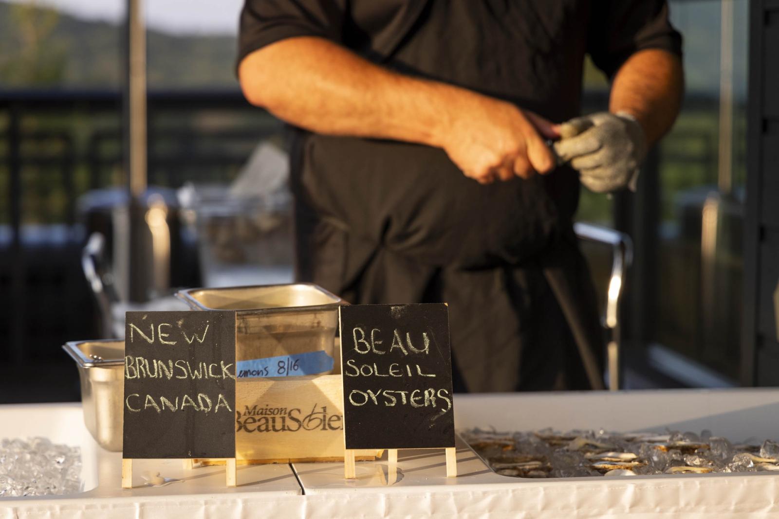 Man shucking oysters at a table with black aprons and gloves.
