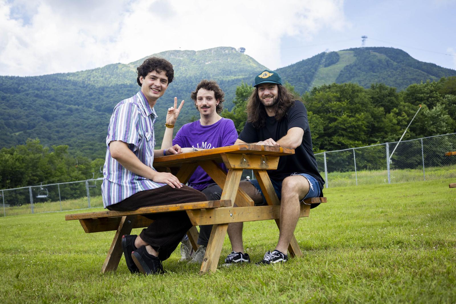 Three people sitting at a picnic table on grassy field with mountains in the background.