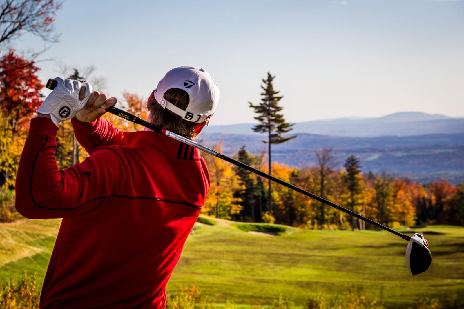 Golfer in red swings club on an autumn course, trees in vibrant colors.