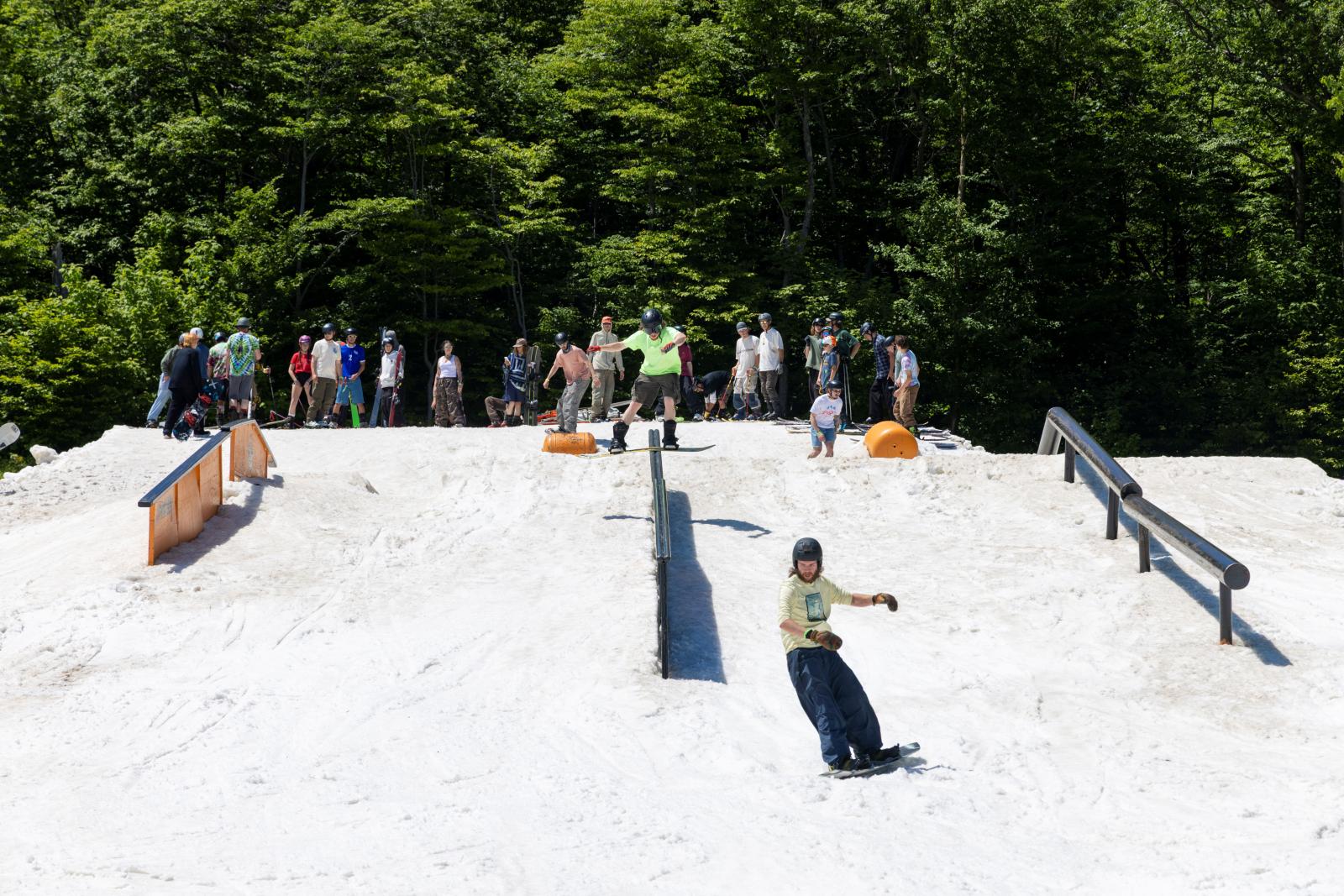 Snowboarder descends a snowy hill, surrounded by spectators and trees in the background.