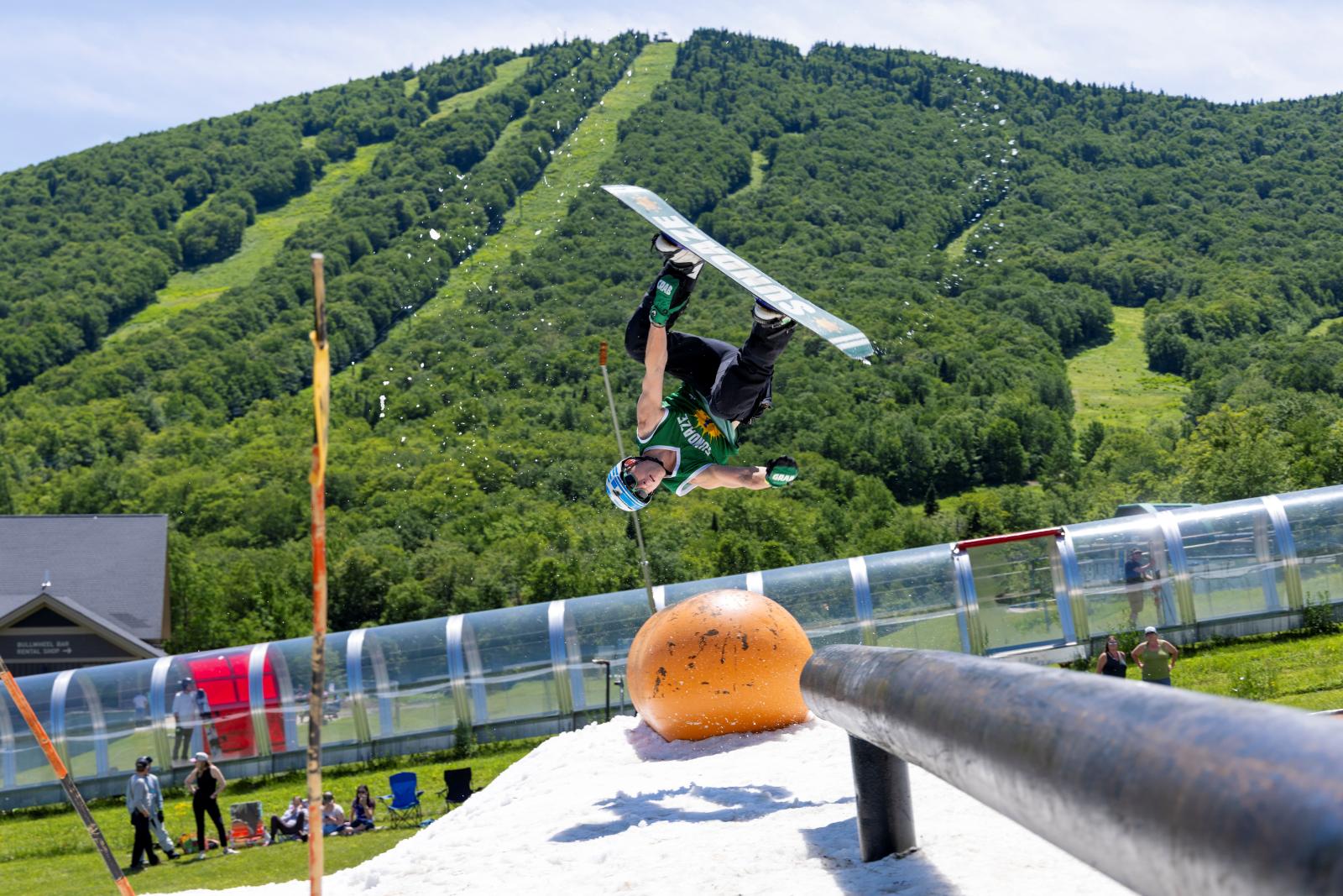 Snowboarder mid-air trick on a summer slope, green hills in the background.