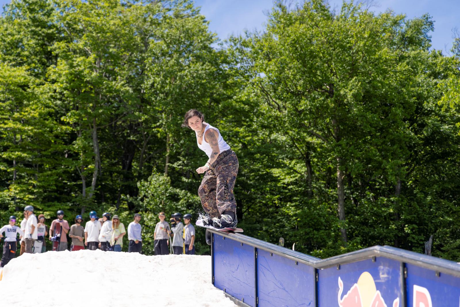 Snowboarder grinding rail, people watching, green trees, blue sky.
