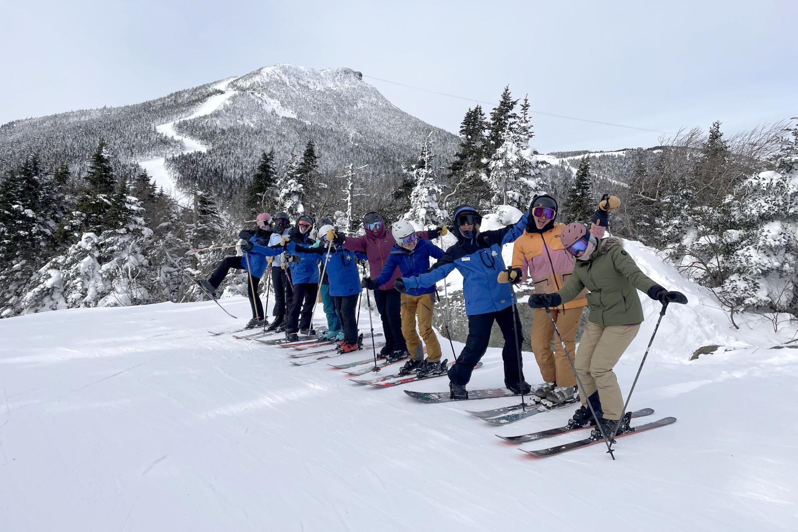Female skiers and snowboarders in colorful gear pose on a snowy slope with a mountain backdrop.
