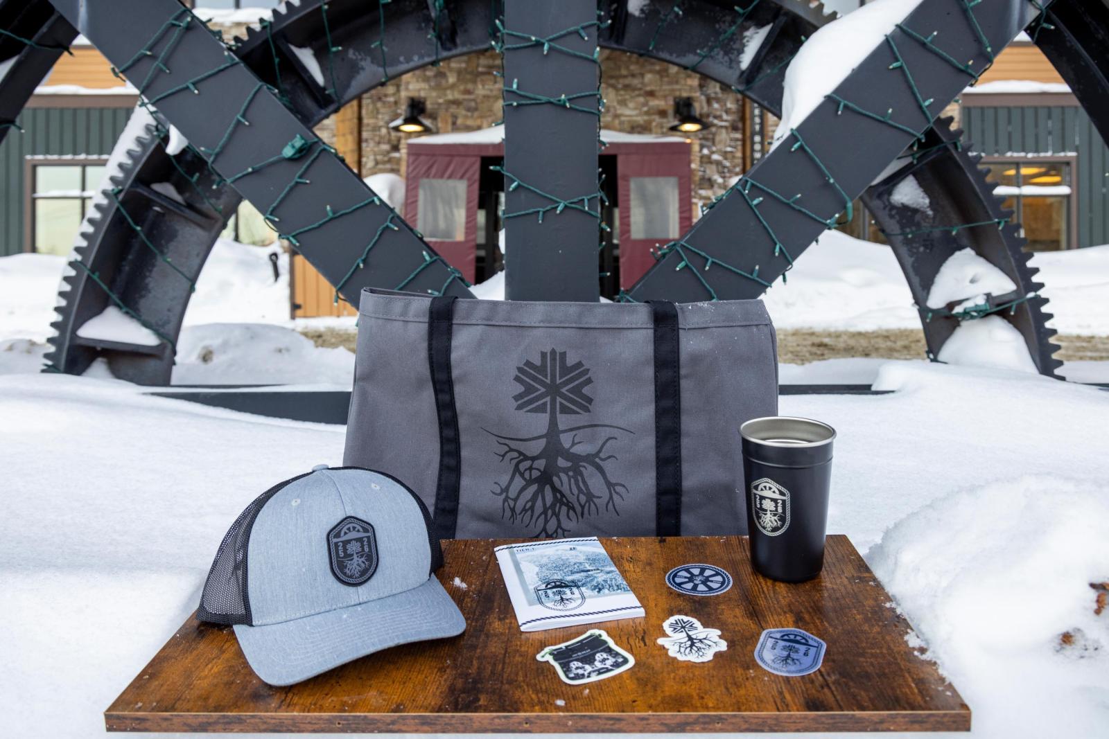 Hat, tote bag, tumbler, and stickers on a snowy table outside.