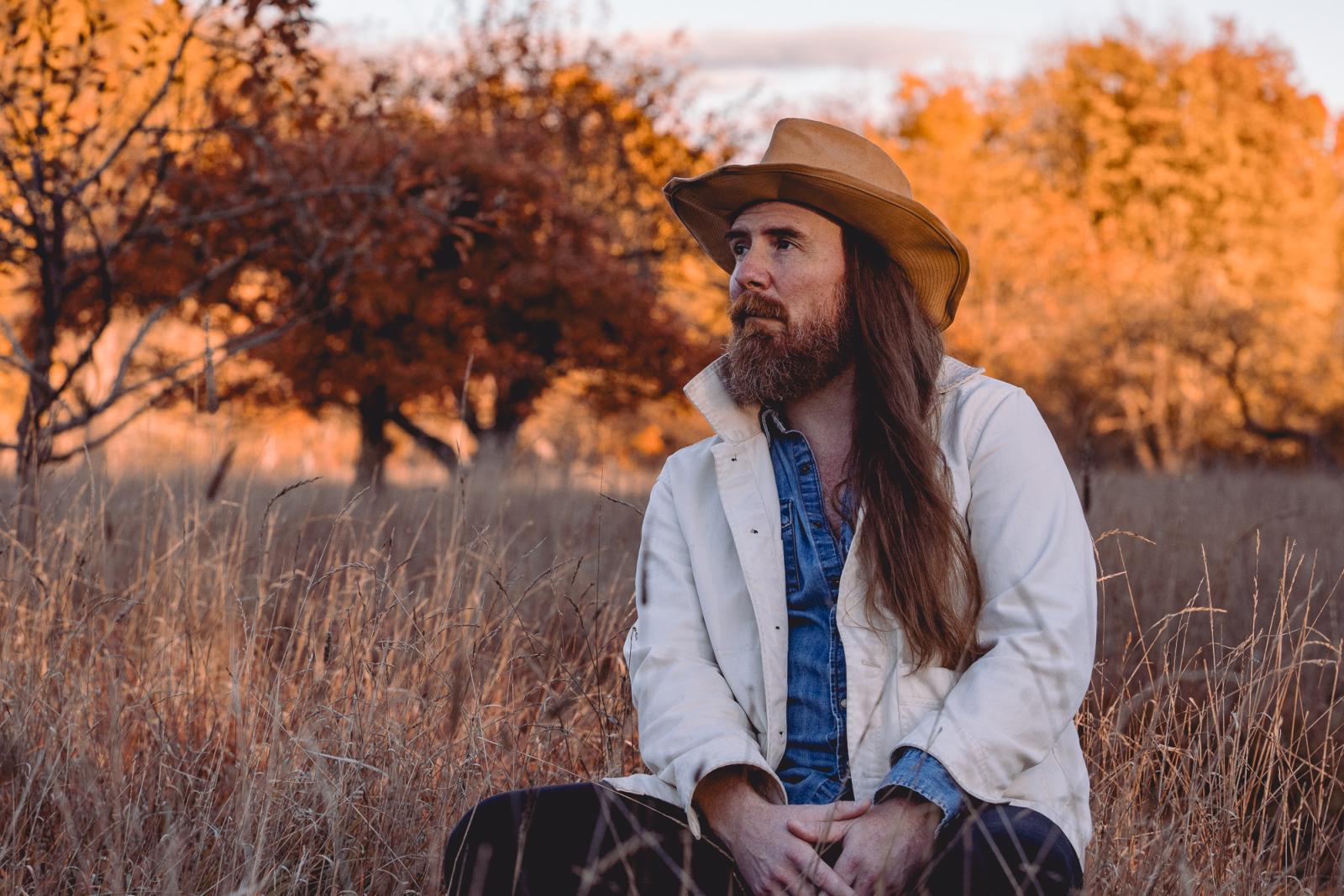 Bob Wagner in a cowboy hat sits in a field with autumn trees.