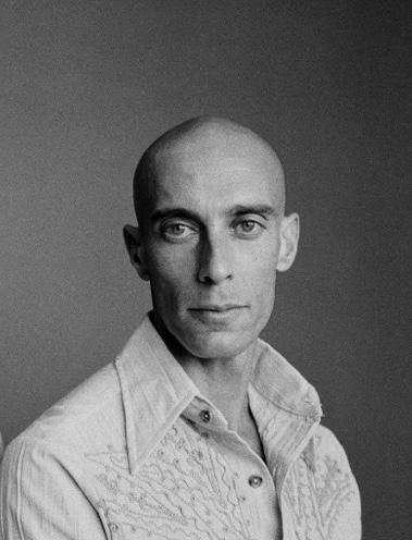 Bald person in a patterned shirt, facing camera, against a gray background.