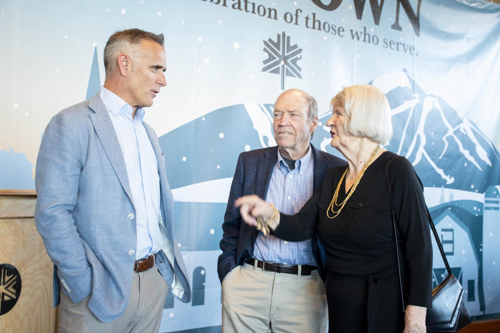 Three people in conversation, two men and one woman, indoors with a snowy mountain backdrop.