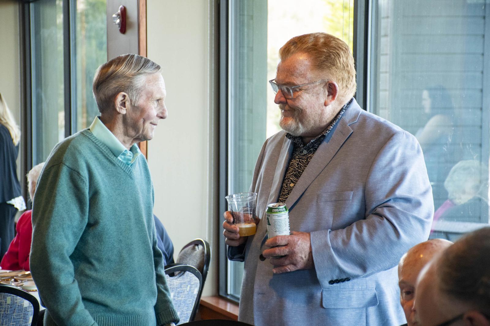 Two men in conversation, smiling, holding drinks in a bright room.