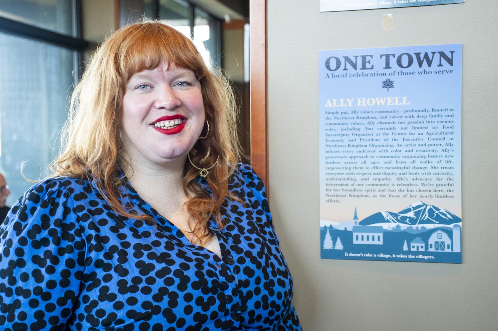 Smiling woman with red hair, in blue leopard-print top, standing beside a blue poster.