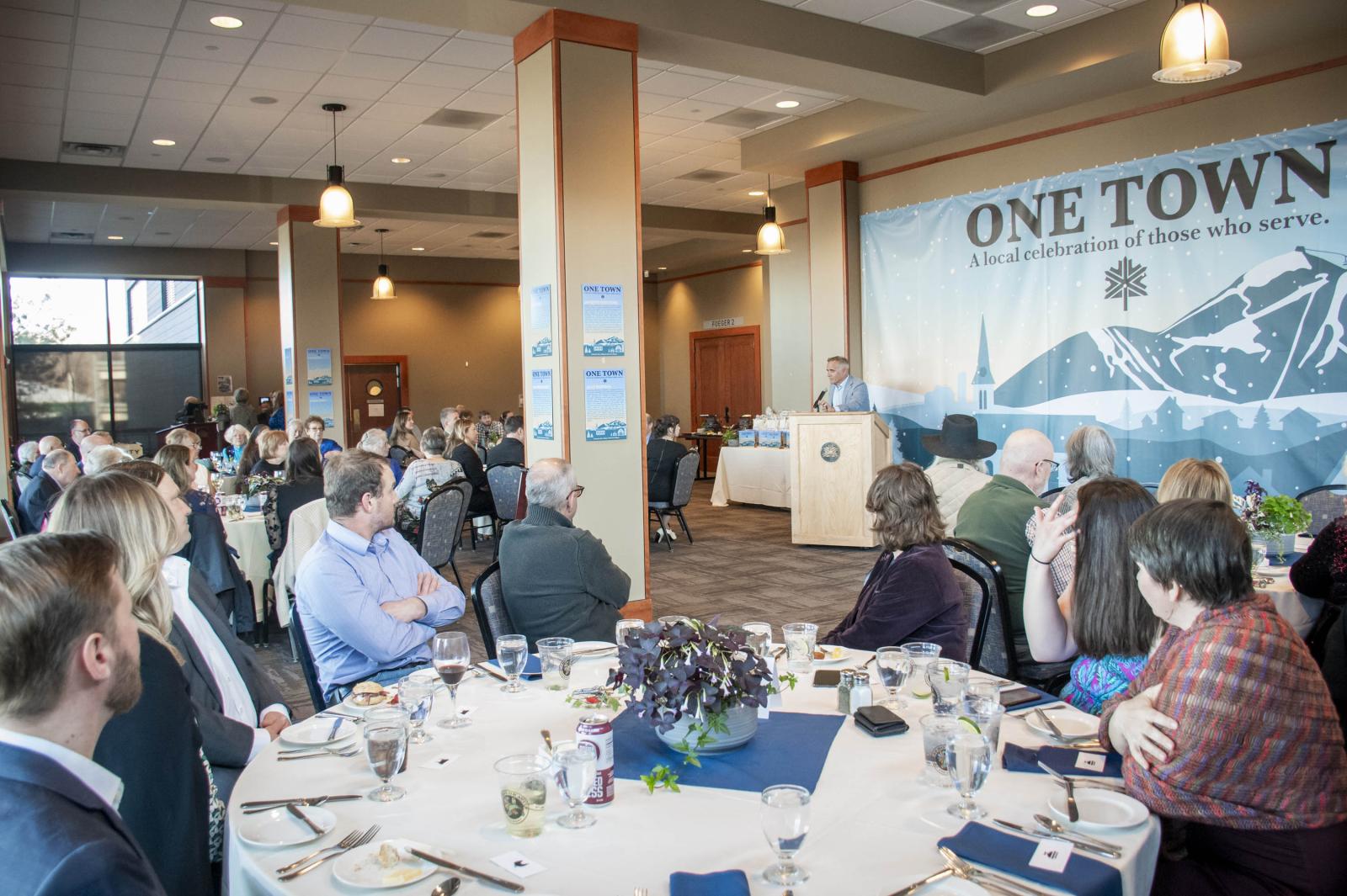 Conference room with people seated around tables, speaker at podium, banner in background.
