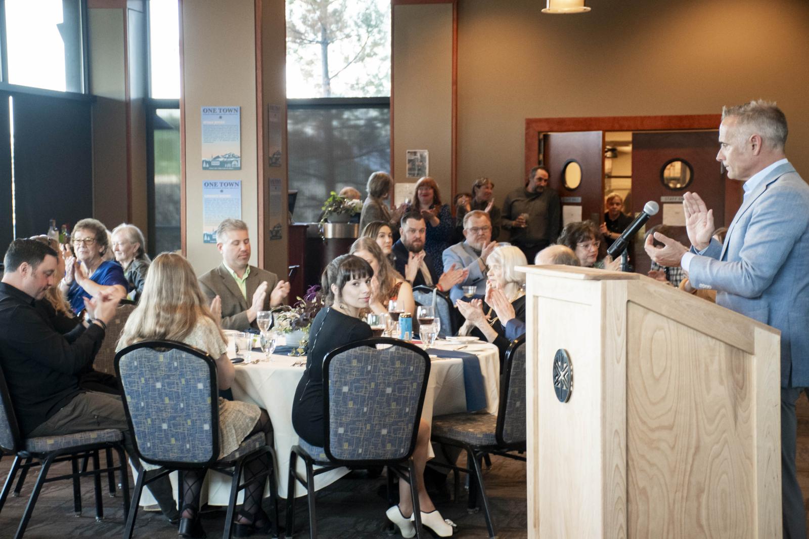 Audience claps as a speaker in a suit addresses a dining hall gathering.