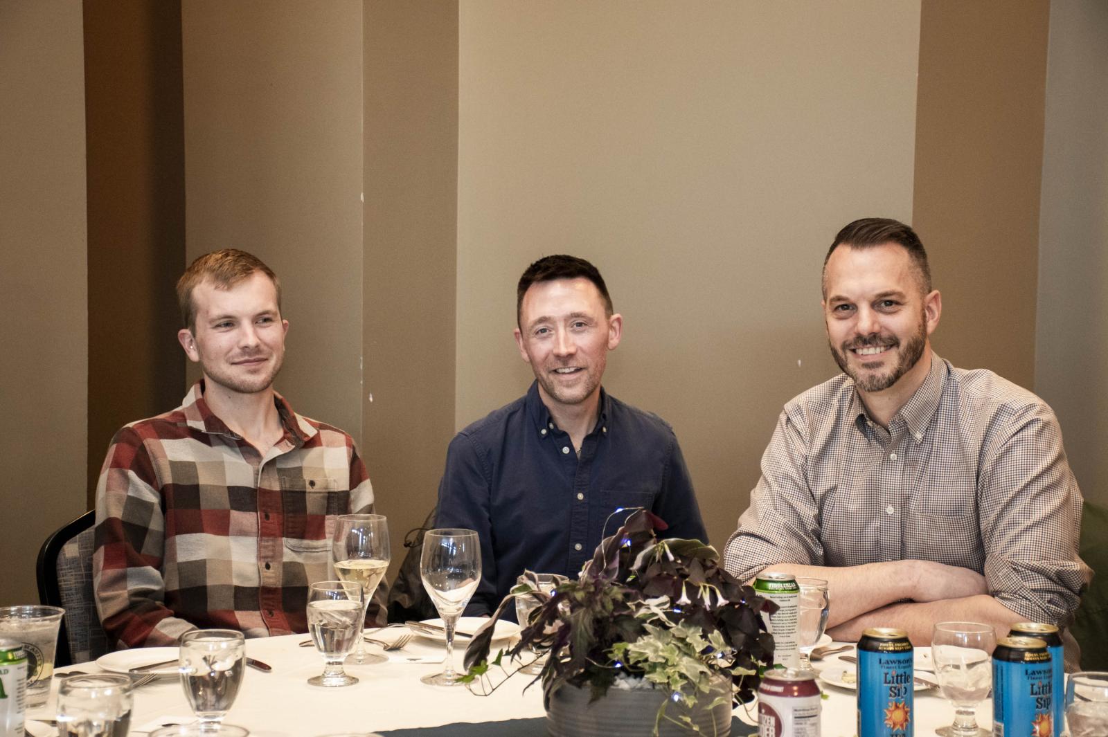 Three men sitting at a round table with drinks and a plant centerpiece.