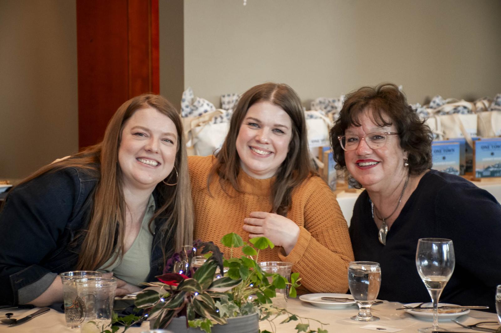 Three women smiling at a table with plants and glasses.